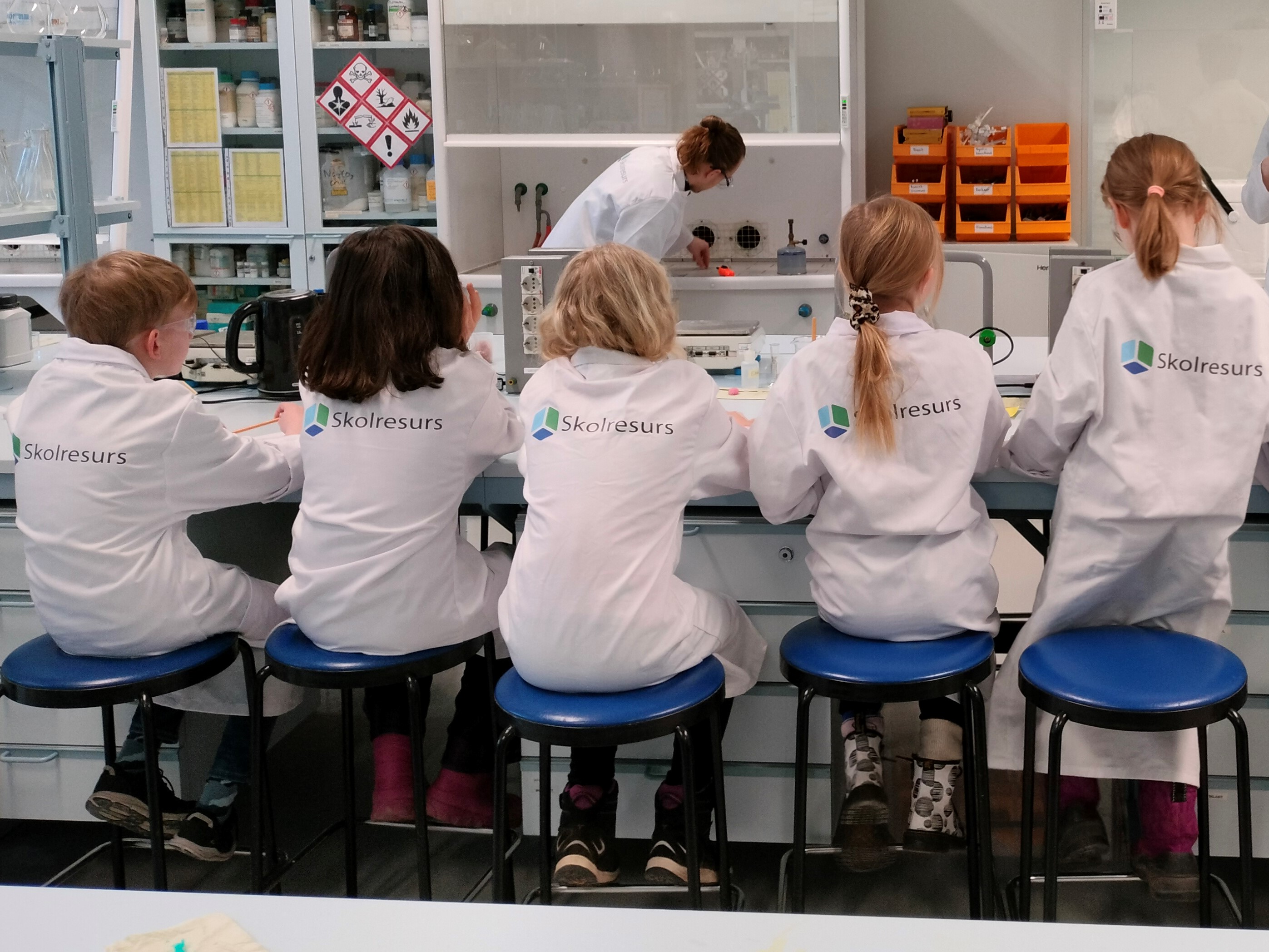 Barn i vita labbrockar sitter på rad vid ett bord med ryggarna mot fotografen. På labbrockarna står det Skolresurs.