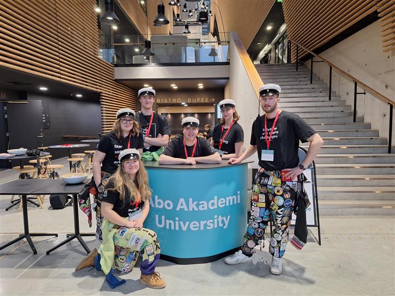 Studerande står vid ett Åbo Akademi bord och ler mot kameran under DI-dagen 2026. 